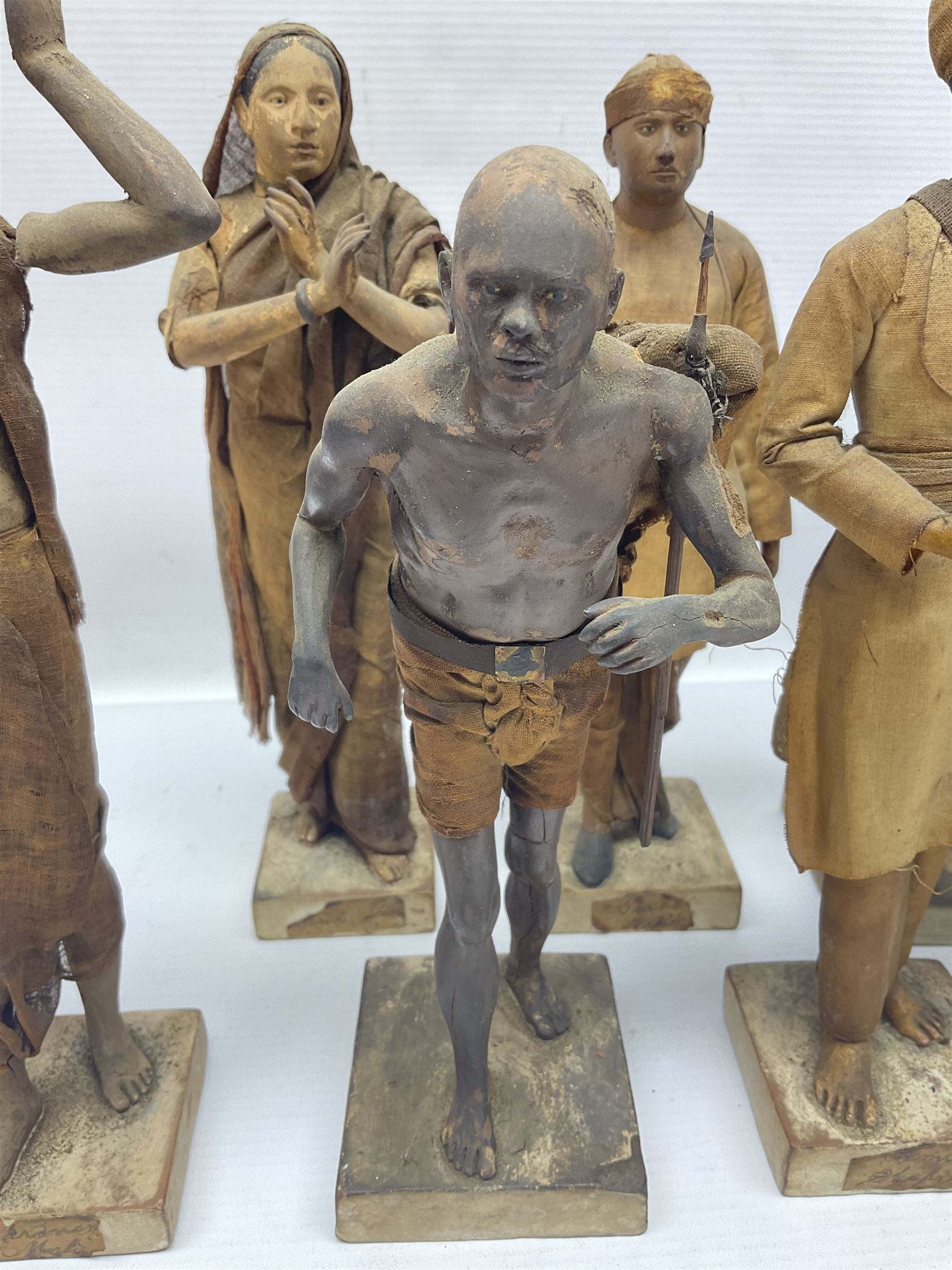 Seven 19th Century Indian clay dolls, possibly Krishnanagar, each figure with painted details, wearing cloth garments, standing on rectangular bases, some with paper label to the base, H25cm