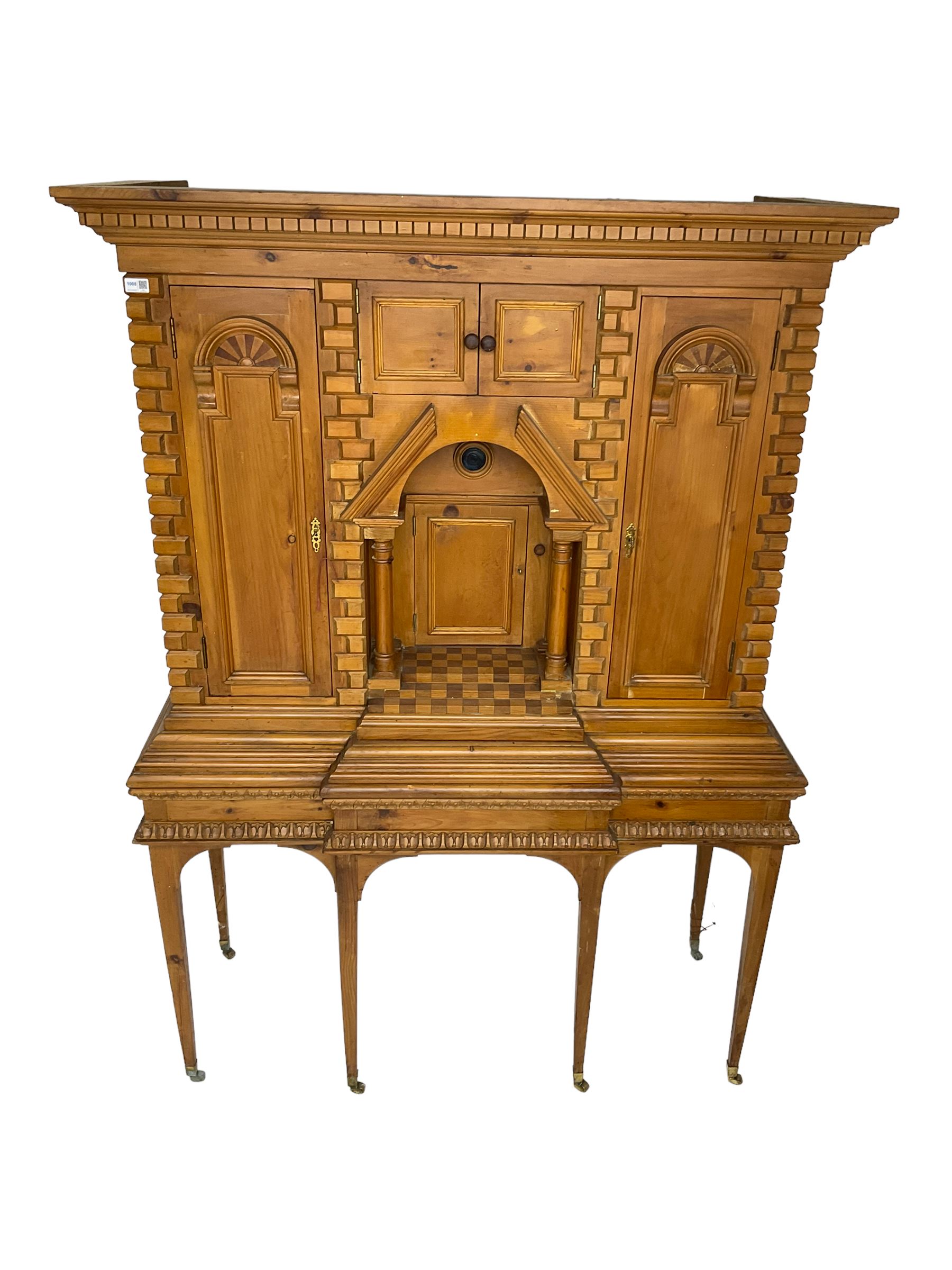 20th century pine architectural collector's cabinet on stand, projecting dentil cornice over various compartments, central recessed arch with turned pillar supports and sloped arch pediment, fitted with four cupboards, false brickwork uprights to front and corners, central hidden drawer revealing cube work surface, lower moulded plinth, the stand with foliate carved mouldings, on six square tapering supports with brass castors