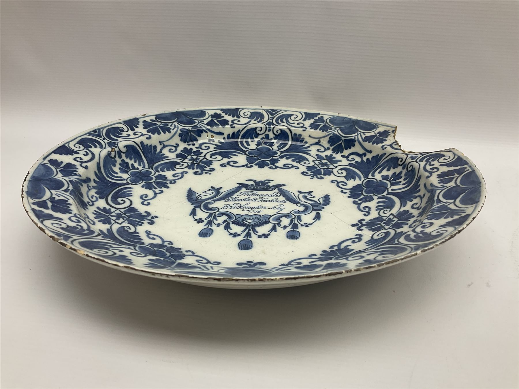Early 18th century Delft marriage plate, of circular form with dished centre, the centre inscribed and dated 'Thomas and Elizabeth Kockerill Bridlington Key 1715' within a crowned C scroll cartouche, flanked by demi-griffins, above a putto head and tassels, within a wide foliate border, D34cm
