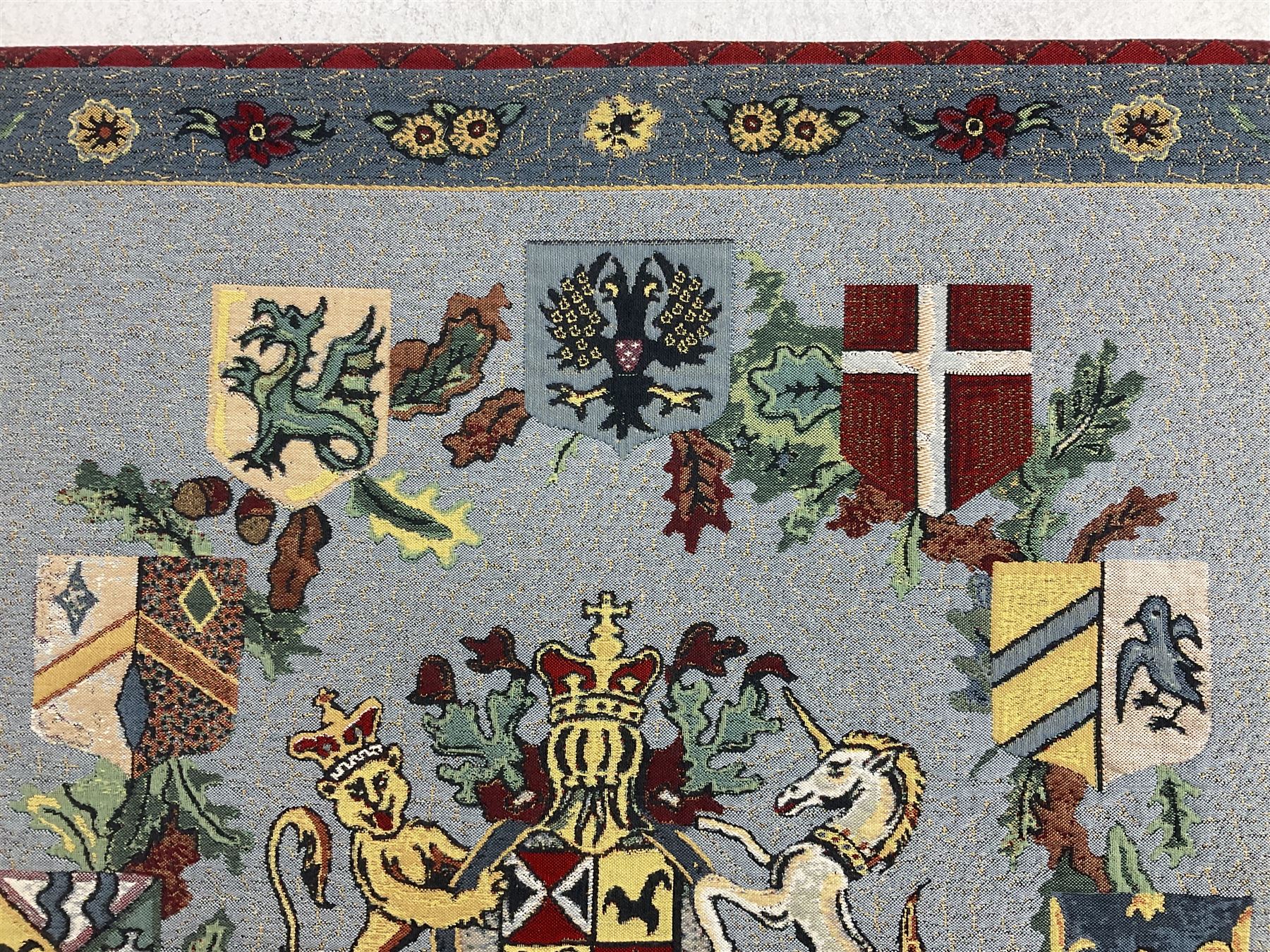 Three tapestries comprising 20th century wool tapestry panel of a medieval nobleman upon a horse with a castle in the distance, together with a smaller tapestry depicting The English Coat of Arms, both hung on rails, and further depicting village scene, largest approx 132cm x 95cm