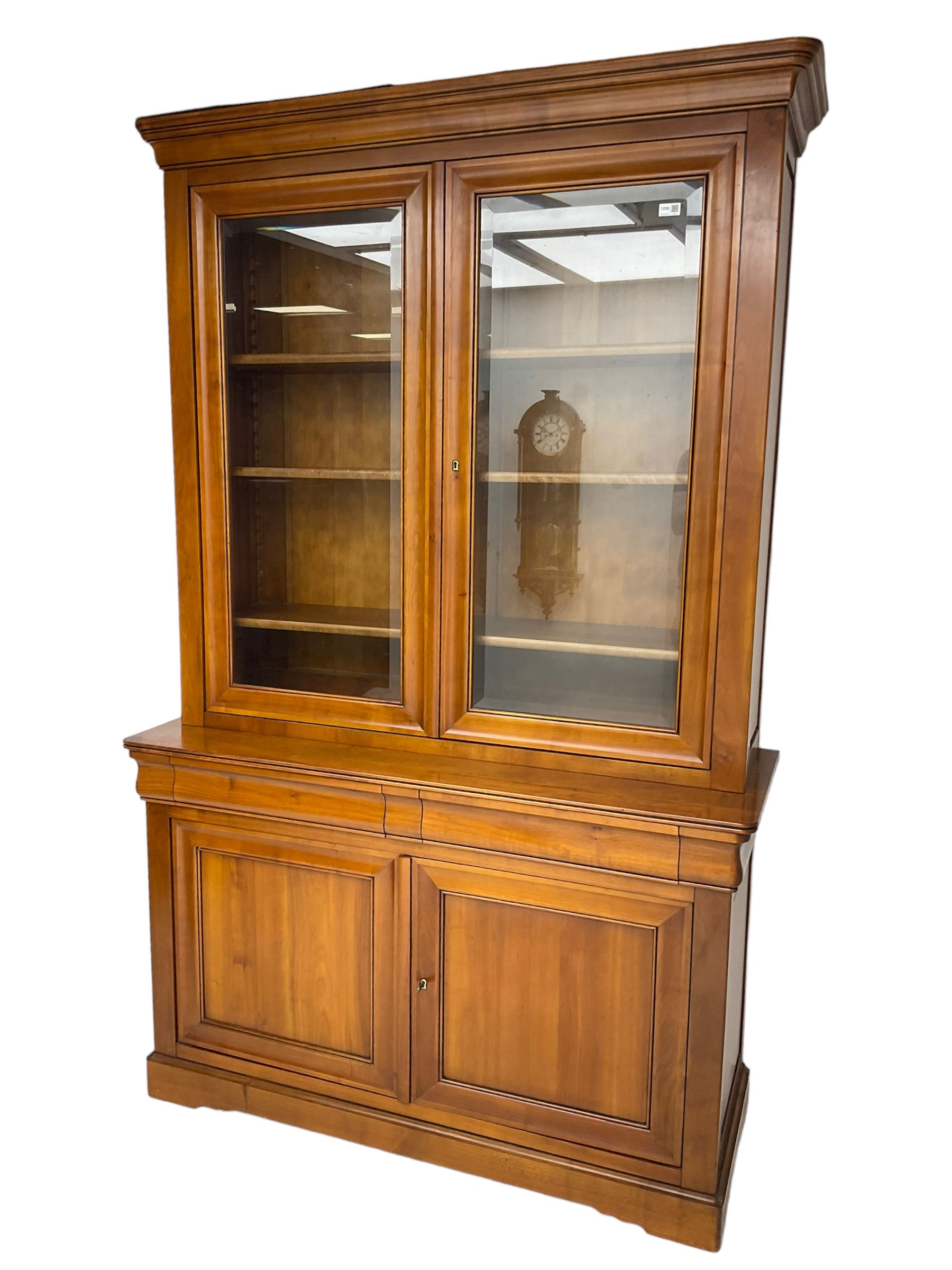 French cherry wood bookcase on cupboard, projecting cornice over two cupboard doors with bevelled glass panels enclosing three adjustable shelves, base fitted with two cushion drawers over double cupboard
