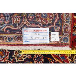 Central Persian Kashan hand-knotted rug, madder red field, with central lobed medallion with pendants, issuing an all-over design of scrolling vine, palmettes, and floral sprays, with conforming spandrels, enclosed by a broad indigo palmette and vine border framed by guard stripes in a palette of navy, blue and ivory tones, with cotton fringe ends 