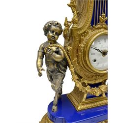 A 20th century continental gilt metal Lyre mantle clock with a pair of matching six light candelabra, spring driven movement housed in a gilt drum case on a brass and cobalt blue base with paw feet, two bronzed metal figures of mythological cherubs with animal legs and cloven hooves supporting a festooned garland swag, eight-day twin barrel striking movement with a floating lever balance escapement, striking the hours and half-hours on two bells, white enamel dial with roman numerals, minute track and pierced steel hands, dial inscribed “Imperial”. 