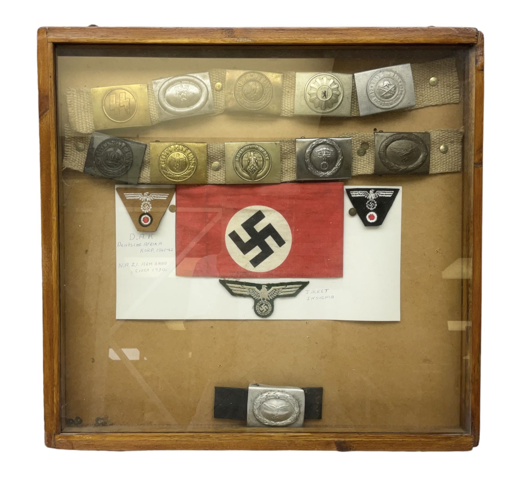 Display of eleven German belt buckles including 'SS', Party Buckle, two Army, Fire Service, two Luftwaffe, NSKK, Hitler Youth and WW1 Army; framed with three cloth badges and an armband