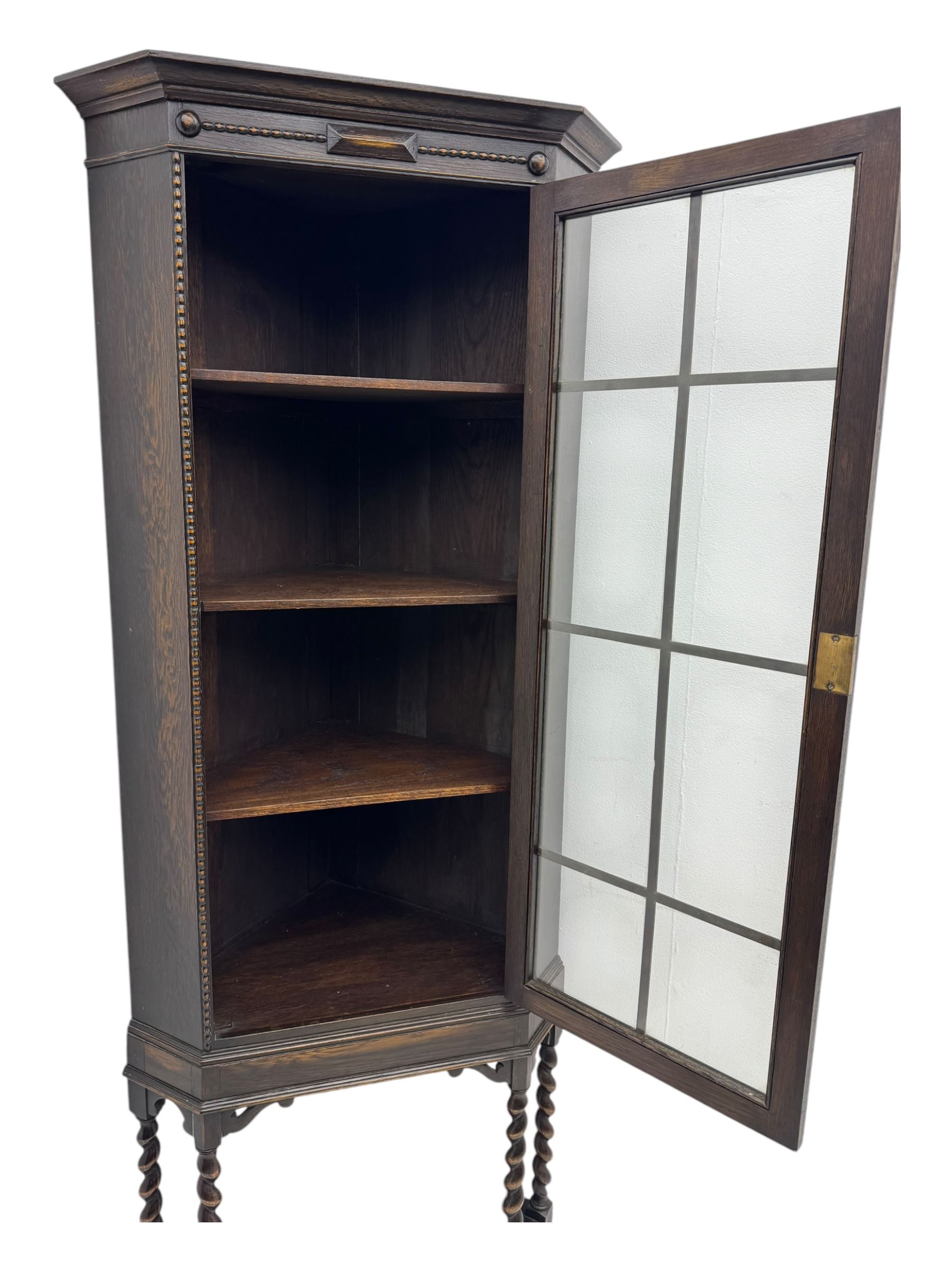 Early 20th century oak corner display cabinet, the moulded canted cornice over a single glazed door with geometric astragal bars, shelved interior, the lower tier forming a triangular platform, raised on four barley-twist supports united by shaped brackets