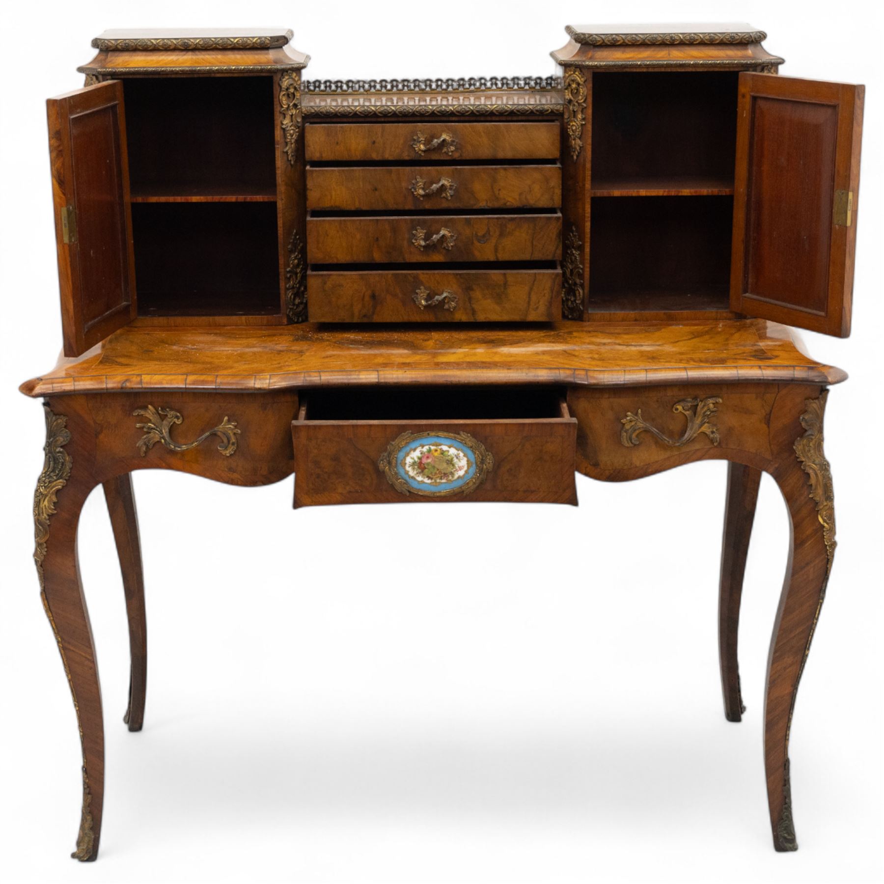 Late 19th-century Louis XV revival gilt bronze mounted and walnut Bonheur-de-Jour, rectangular-shaped serpentine form, with gilt bronze beading, frieze with two blind drawers and gilt bronze handles, centre drawer with large central serves style porcelain plaque, the elevated section with brass gallery above panelled cupboard doors also with serves style porcelain plaques and gilt bronze mounts, interior with single shelf, central section with four drawers each with gilt bronze handles, raised on slender cabriole legs fitted with gilt bronze rococo style masks and sabots