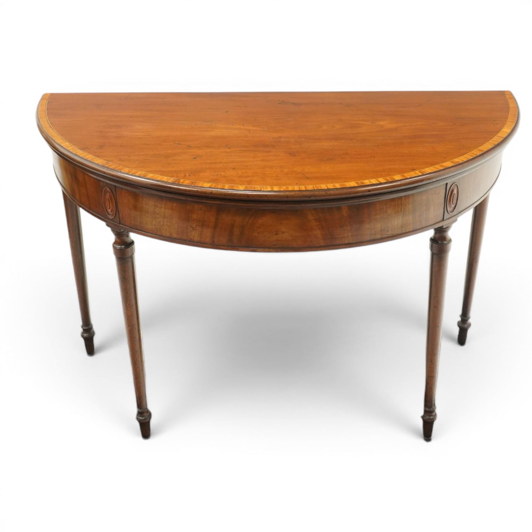 Georgian mahogany demi-lune card table, the lunette shaped top with cross-banded and boxwood-strung edge, opening to reveal baize-lined interior and supported by rear gatelegs, over a banded frieze with applied oval plaques, raised on turned and tapering supports