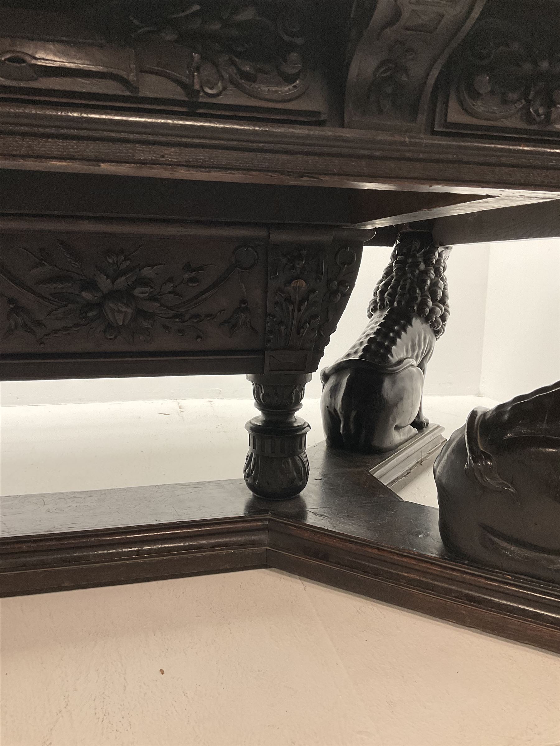 Large 19th century carved oak library table, the rectangular top with lunette carved edge over cartouche panelled and carved  scroll frieze, the central tapering rectangular support relief carved with masks and garlands, on four carved seated lion supports at each corner, on flat angular stretcher with recessed castors stamped ‘Bartholomew & Fletcher, London’