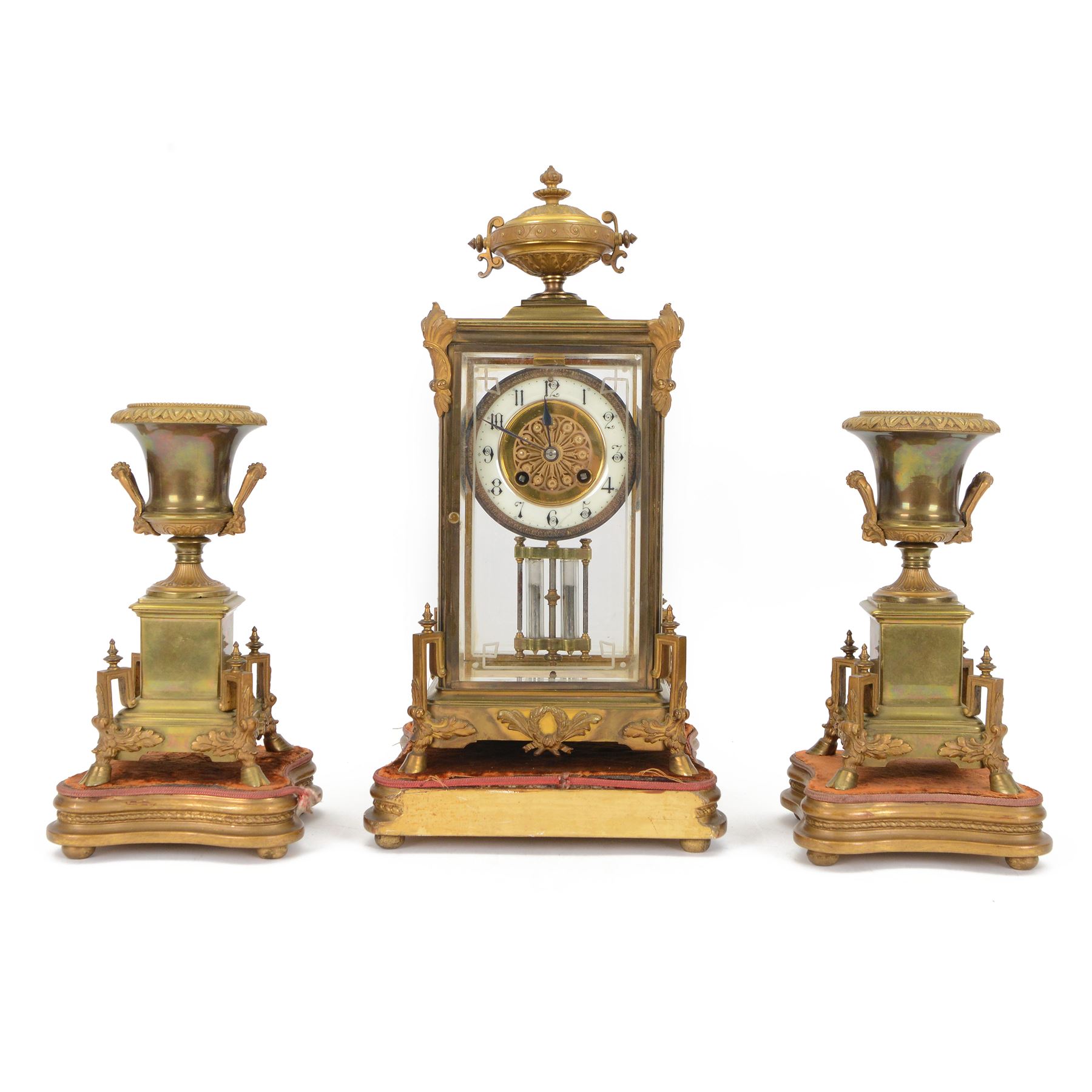 French- Art-deco Edwardian 8-day four-glass clock garniture, with etched glass panels, cast brass decoration to the corners of the case and splayed feet representing cloven hoofs to the base, case surmounted by a large oval urn on a tiered brass platform, two part dial with a recessed brass centre comprising of pierced radial decoration, with steel spade hands and Arabic numerals to the chapter, rack striking movement with a deadbeat Brocot escapement, striking the hours and half-hours on a bell. With a visible twin file mercury pendulum. Garni comprising two large twin handled urns on rectangular pedestals raised on conforming feet representing cloven hoofs, Clock and garni presented on three shaped wooden gesso bases with velvet pads. With key.