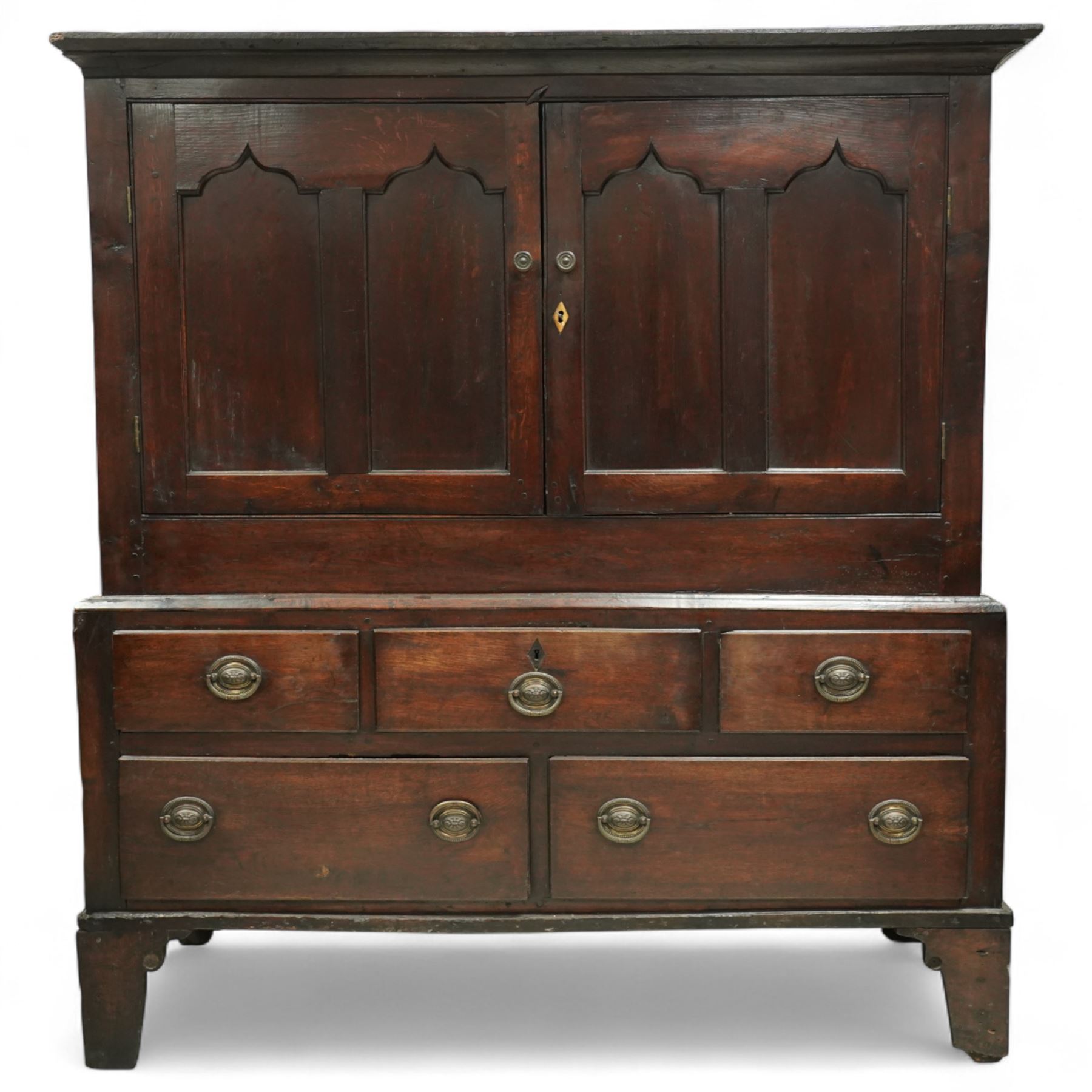 18th century oak dwarf press cupboard, the upper section with twin arched panel doors above three short and two long drawers, each with brass plate handles, shaped apron and stile feet, of compact country form