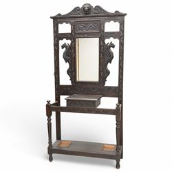 Late Victorian heavily carved oak hall stand, the scrollwork pediment with carved lion's head, bevelled mirror plate flanked by carved dragon motifs, over hinged storage compartment and umbrella stands with drip trays, on turned front supports
