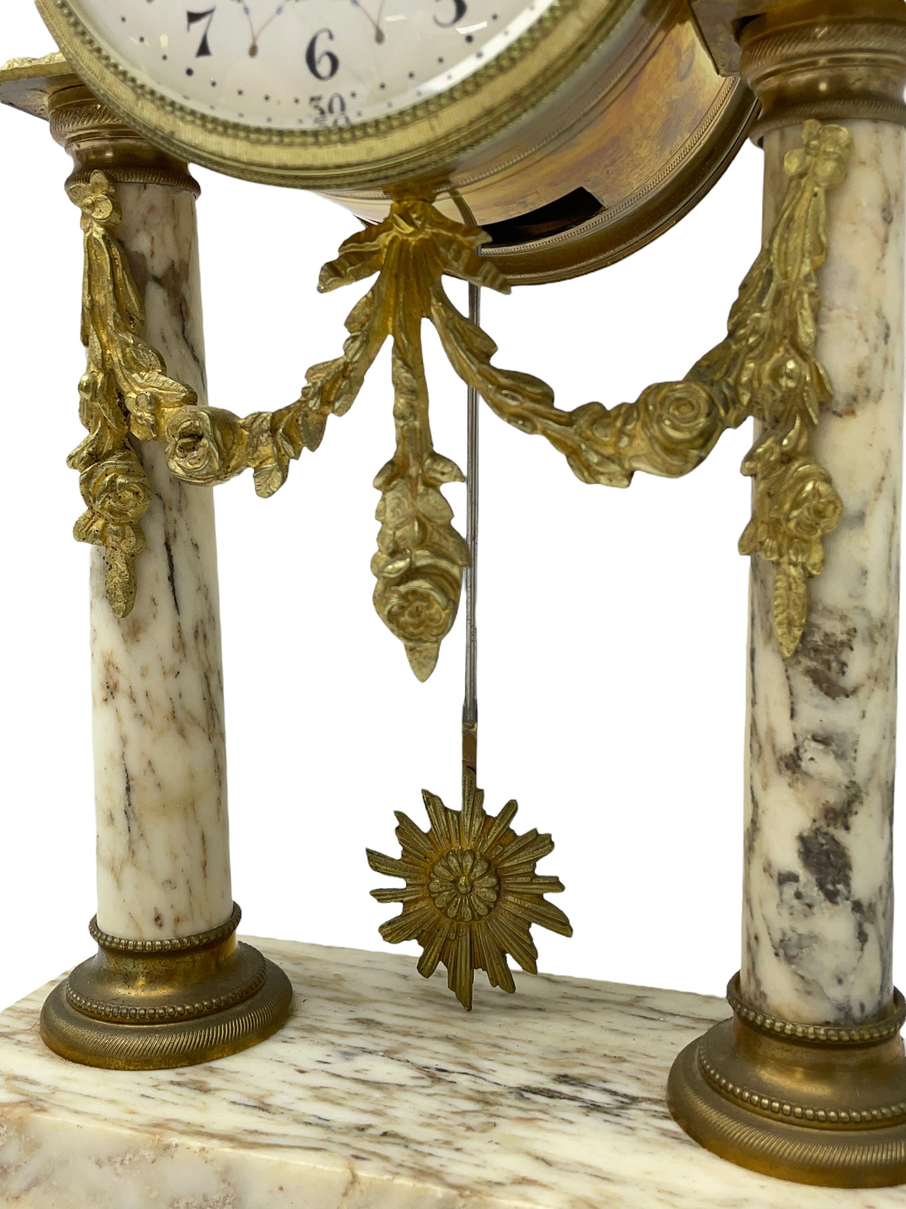 20th century French portico clock c1910 on a white veined marble base, gilt drum movement case supported between two marble columns with brass capitals and a suspended garland swag with a starburst pendulum beneath, enamel dial with Arabic numerals, floral decoration and Louis XV pierced gilt hands, convex glass and brass bezel, 8-day countwheel striking movement striking the hours and half hours on a bell. 




