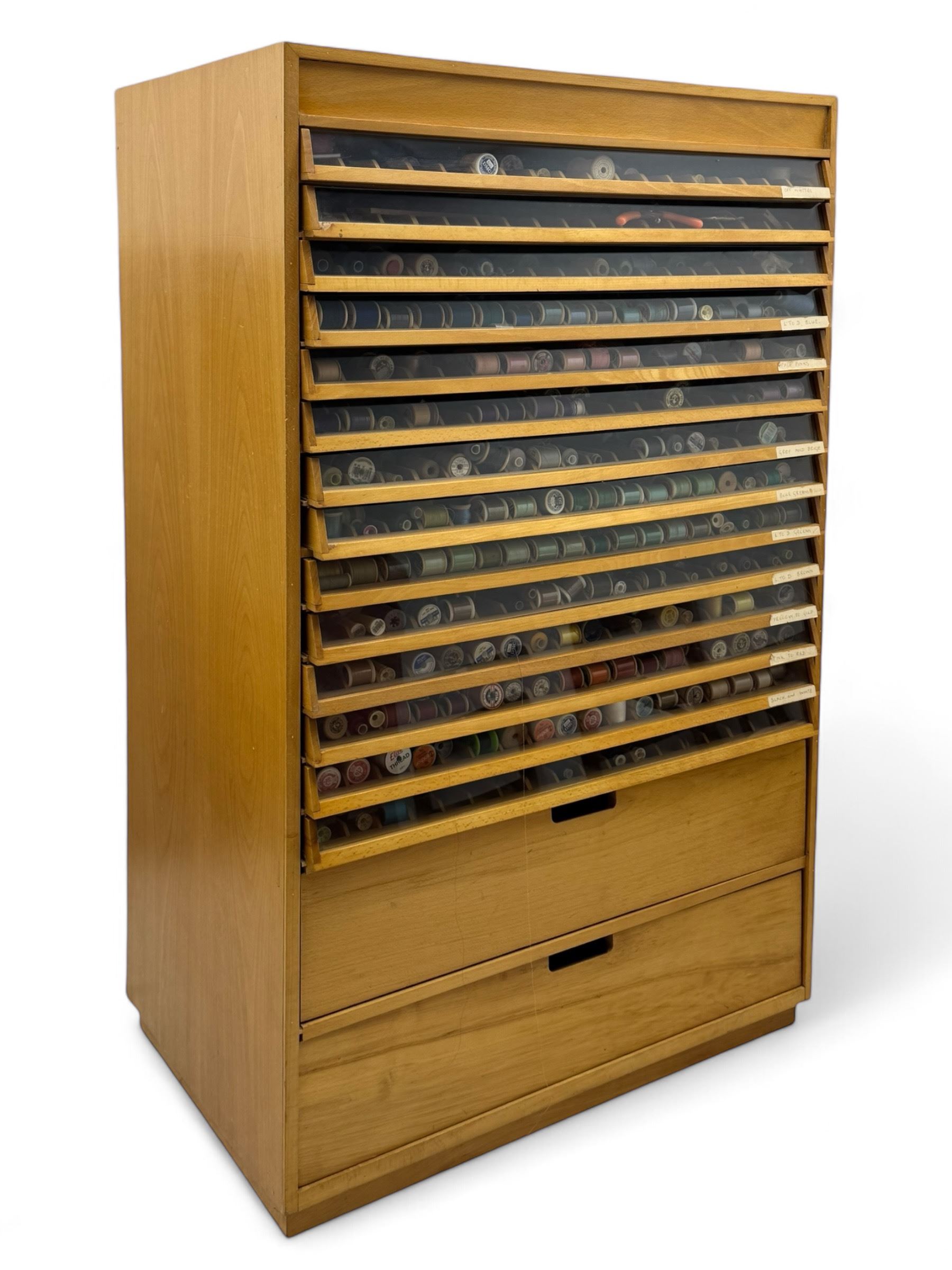 Large beech haberdashery thread storage cabinet, rectangular form with fourteen glass-fronted sloping drawers housing an extensive array of thread spools in assorted colours, over two larger drawers, raised on plinth base