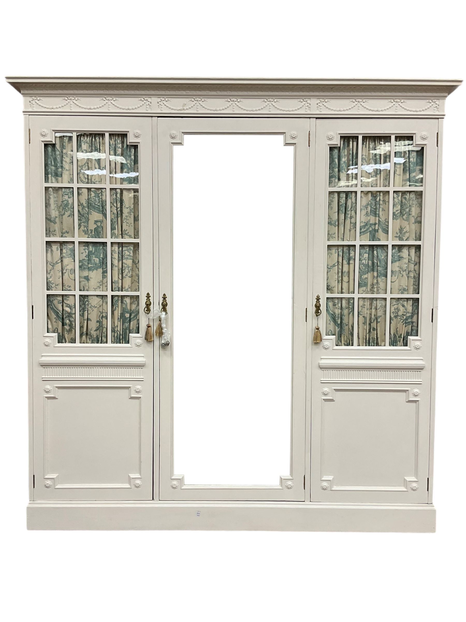 Maple and Co - Edwardian Hepplewhite design white painted triple wardrobe, foliate carved projecting cornice over ribbon and husk swag decorated frieze, two curtain lined astragal glazed doors and centre full mirror door, fitted mahogany interior including slides and drawers, plinth base, label to top of central door