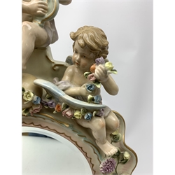  A late 19th century Sitzendorf porcelain mirror, the mirror plate of circular form set within a shaped surround encrusted with flowers and foliage, surmounted by three figures of cherubs and raised upon four scroll feet, with blue cross hatch mark beneath, H57cm.   
