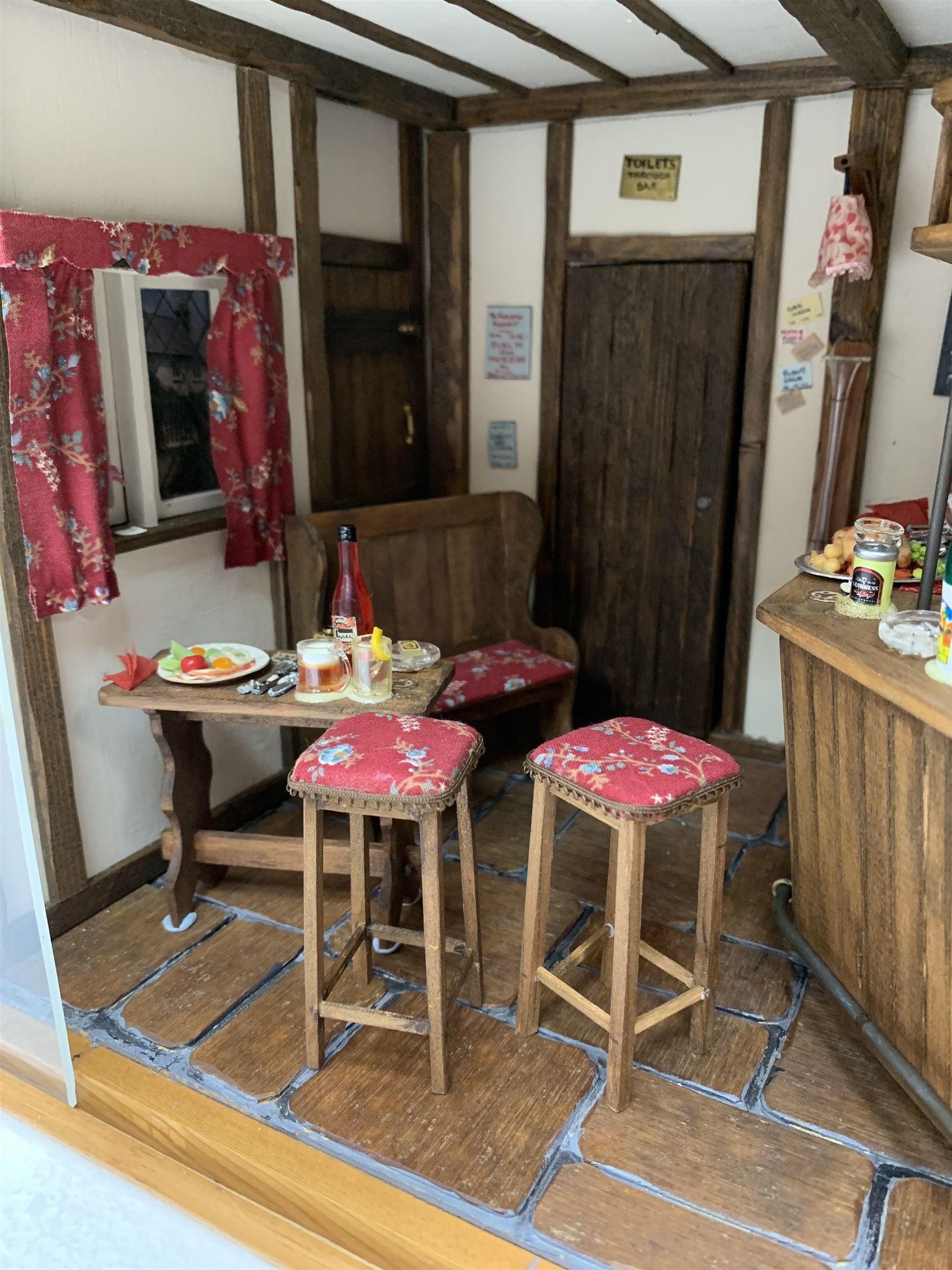 Dolls house scale room setting of a public house bar with tiled floor and beam and plaster ceiling and walls, bench seat, stools and table set out with a meal and drinks, large planked finish canted bar well stocked with food, drinks, bottles, glasses etc and bartender figure behind; electrically illuminated with sliding perspex access panel to front W38cm D28cm H24cm