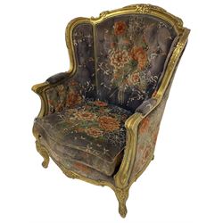 Pair of Louis XVI design gilt framed wingback armchairs, cresting rail carved and moulded with foliate decoration and C-scrolls, upholstered in buttoned floral patterned mauve velvet with loose seat cushion, scallop carved apron over cabriole supports