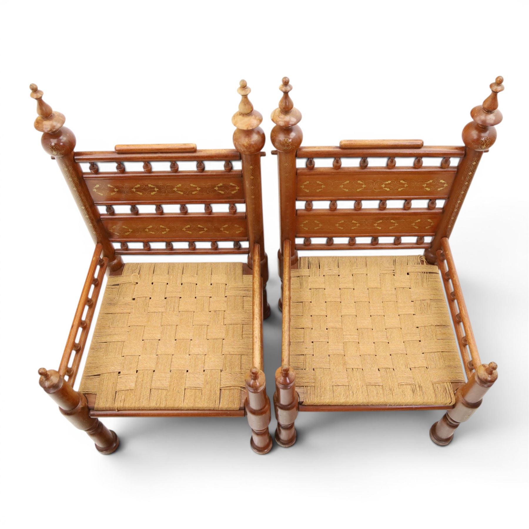 Pair of Anglo-Indian brass inlaid hardwood low chairs, the backs with turned spindle rails and rectangular panels inlaid with scrolling foliate decoration, woven rush seats within moulded frames, raised on turned supports with ring turned feet