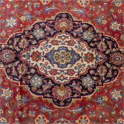 Central Persian Kashan hand-knotted carpet, madder red field with a central cusped medallion of ivory and deep indigo ground, enclosing a stylized floral rosette, framed by scrolling arabesques, field richly ornamented with an intricate network of flowering vinery, palmettes, and delicate floral sprays, spandrels in deep indigo, principal border of navy ground decorated with a continuous palmette and vine-scroll, flanked by multiple guard stripes, with cotton fringe ends 