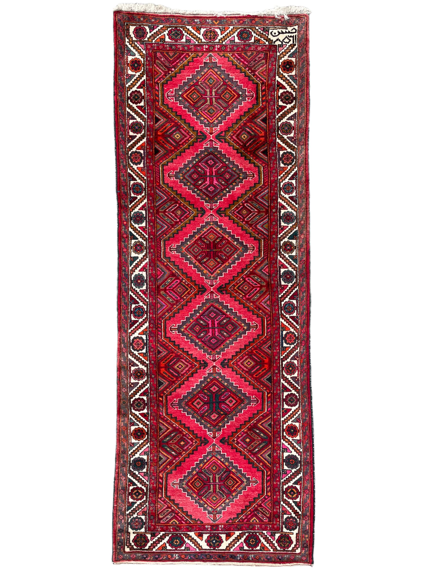 Persian Hamadan red ground rug, decorated with six stepped lozenge medallions, the guarded border with repeating stylised flower head decoration, with inscription to corner
