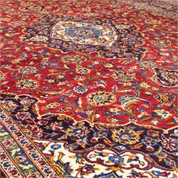 Central Persian Kashan hand-knotted carpet, crimson central field with lobbed medallion in navy blue and ivory ground, surrounded by a dense all-over floral design of scrolling vines, palmettes, and stylized blossoms, enclosed within a deep navy main border, flanked by multiple guard stripes with fringe ends 


