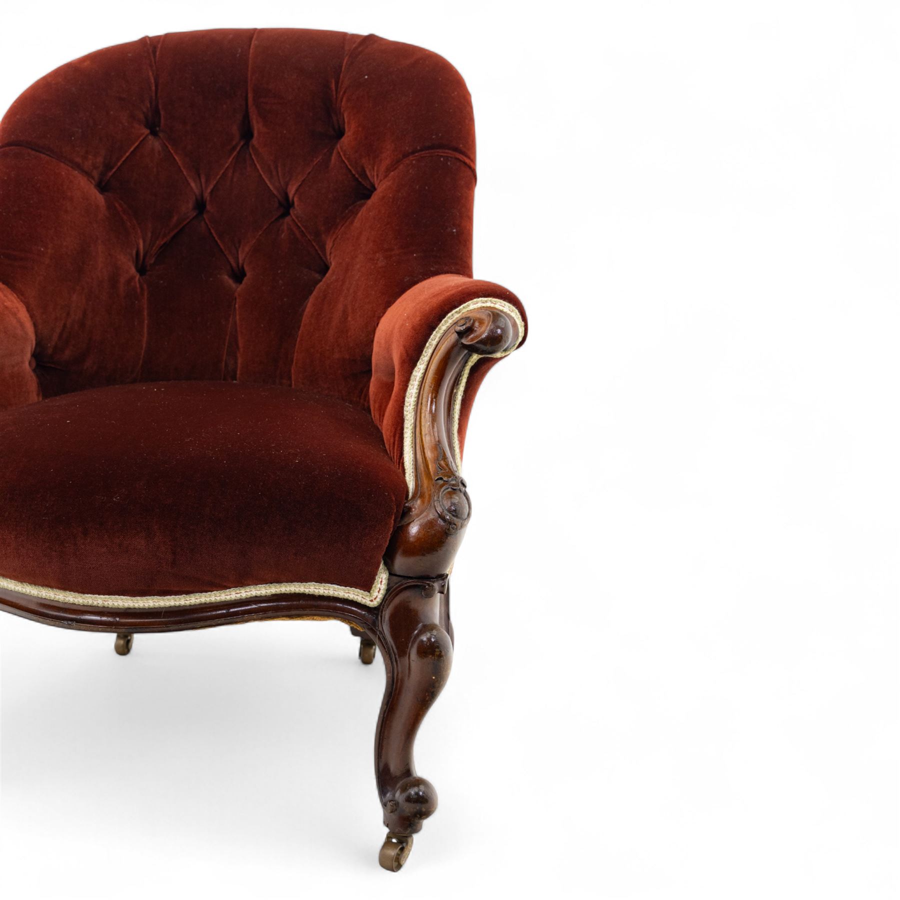 Victorian mahogany framed crimson velvet upholstered tub armchair, with buttoned back and scroll carved arms above a serpentine fronted seat rail, raised upon scroll carved cabriole front legs and plain curved back legs with brass castors 