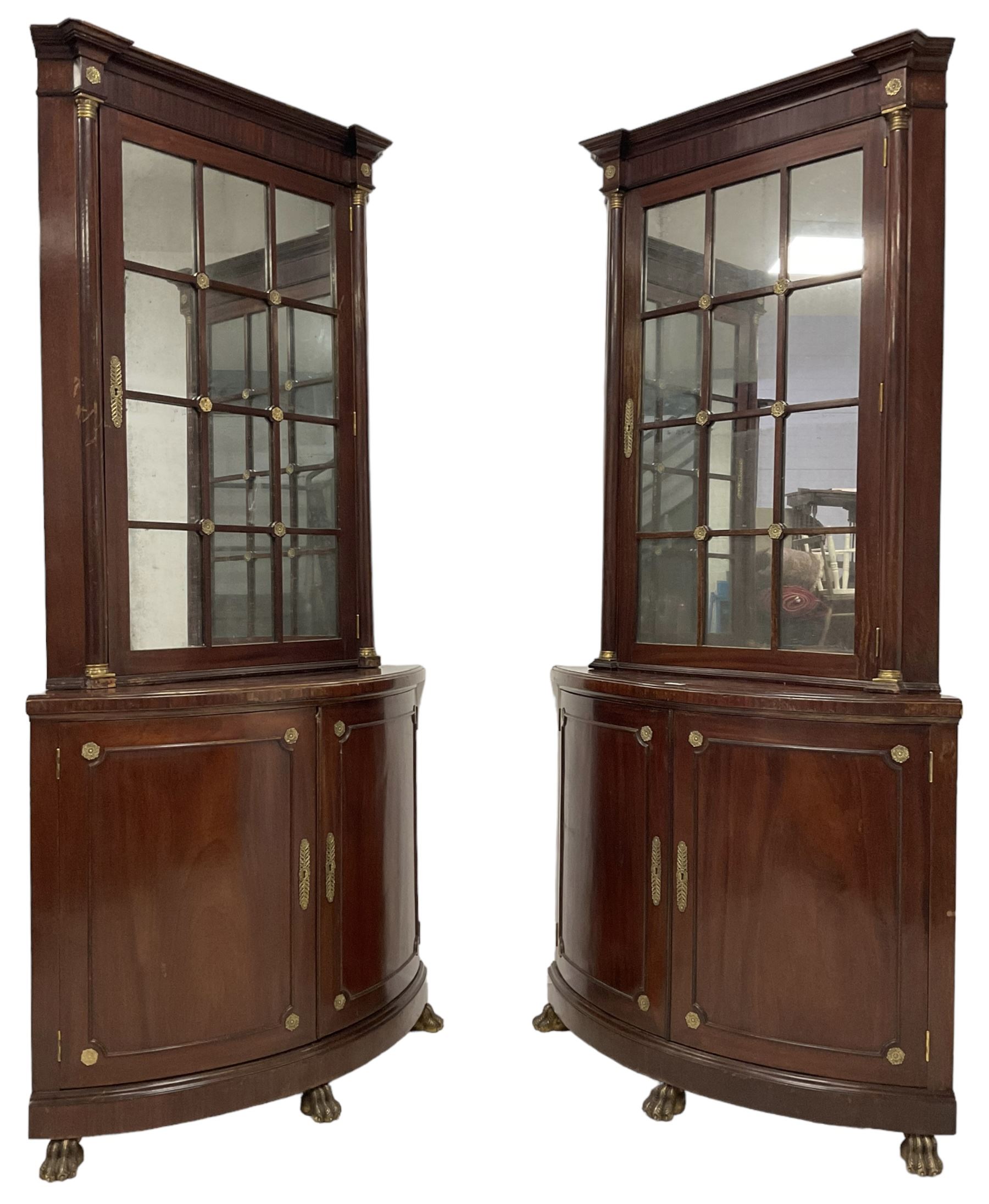 Pair of French Empire design corner cabinets, the straight-front upper section with canted corners and applied half-column pilasters, enclosed by mirror glazed door, the bow-front lower section enclosed by two doors with applied mouldings, mounted by cast metal floral rosettes, skirted base with cast gilt metal hairy paw brass feet