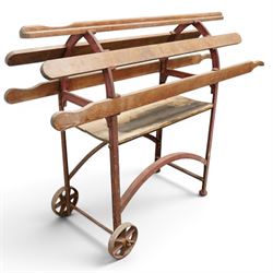 Cast iron and wooden saddle rack, arch form with shelf beneath and wheels to one end, pair of shaped handle poles