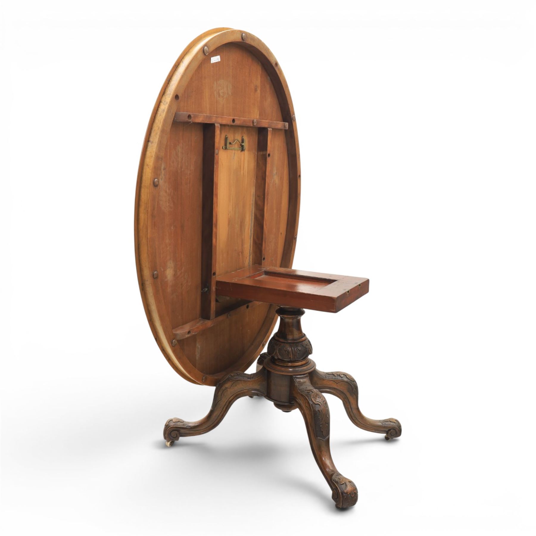 Victorian figured walnut oval loo tilt top table, raised on a heavily carved quatriform base with scrolled supports, terminating in castors