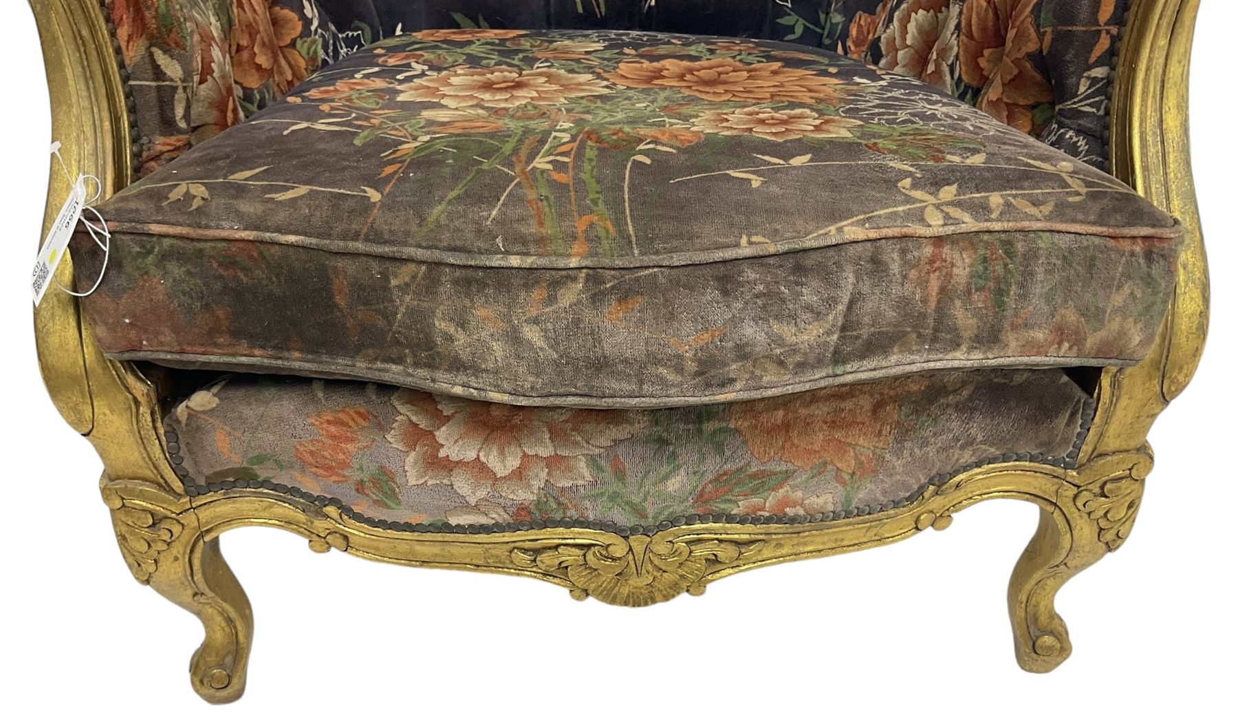 Pair of Louis XVI design gilt framed wingback armchairs, cresting rail carved and moulded with foliate decoration and C-scrolls, upholstered in buttoned floral patterned mauve velvet with loose seat cushion, scallop carved apron over cabriole supports