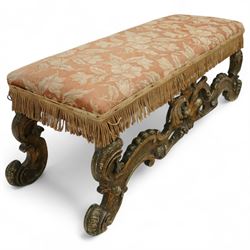 19th-century Rococo-style giltwood window seat, upholstered in floral-patterned coral-ground fabric with fringed border, the intricate scrolled supports united by pierced scrolling foliate stretchers with acanthus and C-scroll decoration