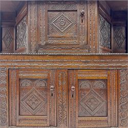 Early 20th-century  oak Jacobean two-tier revival court cupboard, rectangular top with carved notch and stylized foliate carved edge above a carved lozenge pattern frieze and a central cupboard with carved geometric designs and geometric carved panelled sides, above a carved panelled cupboard with two doors of similar geometric design, the whole flanked by two sets of stop-fluted baluster turned columns and supported on stile feet