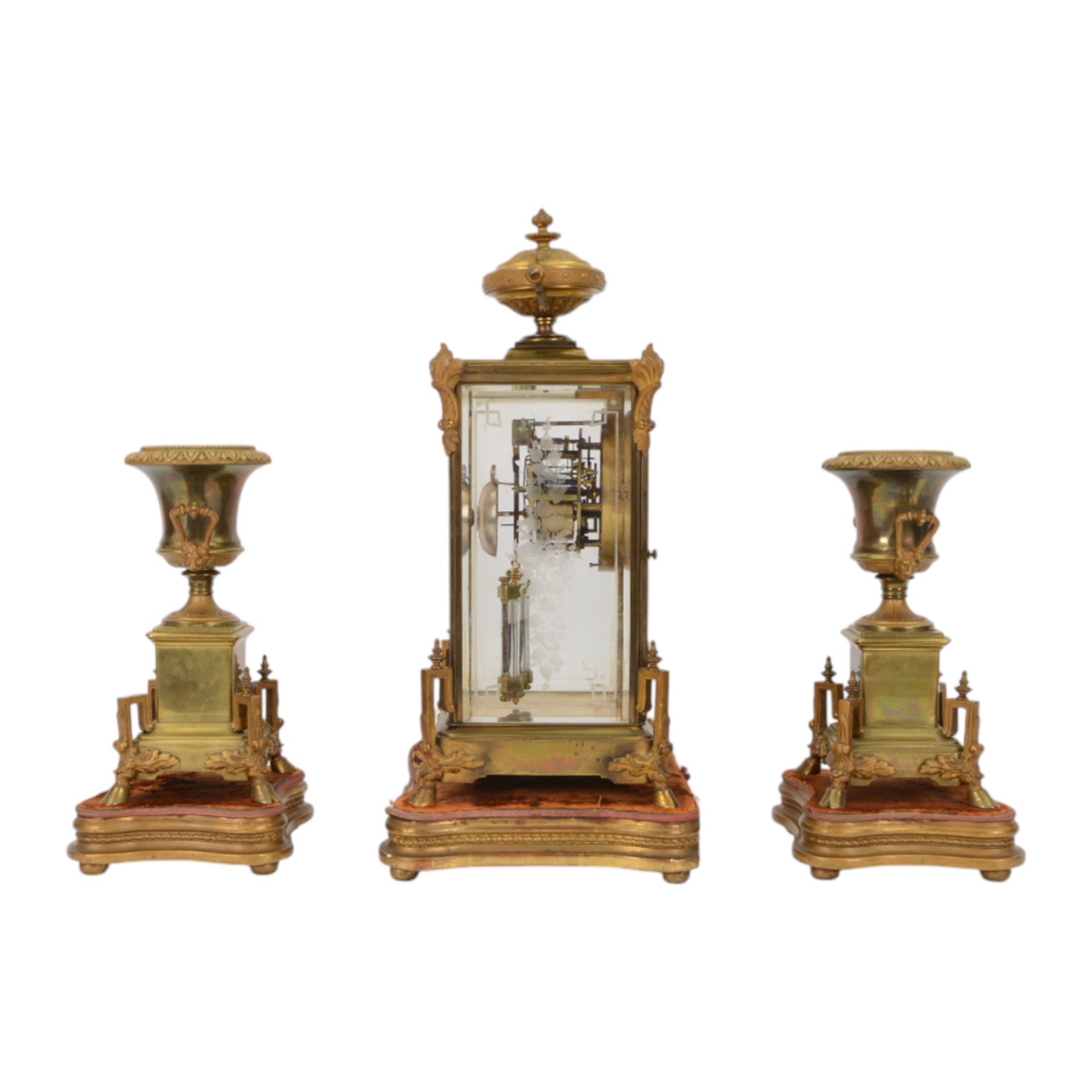 French- Art-deco Edwardian 8-day four-glass clock garniture, with etched glass panels, cast brass decoration to the corners of the case and splayed feet representing cloven hoofs to the base, case surmounted by a large oval urn on a tiered brass platform, two part dial with a recessed brass centre comprising of pierced radial decoration, with steel spade hands and Arabic numerals to the chapter, rack striking movement with a deadbeat Brocot escapement, striking the hours and half-hours on a bell. With a visible twin file mercury pendulum. Garni comprising two large twin handled urns on rectangular pedestals raised on conforming feet representing cloven hoofs, Clock and garni presented on three shaped wooden gesso bases with velvet pads. With key.