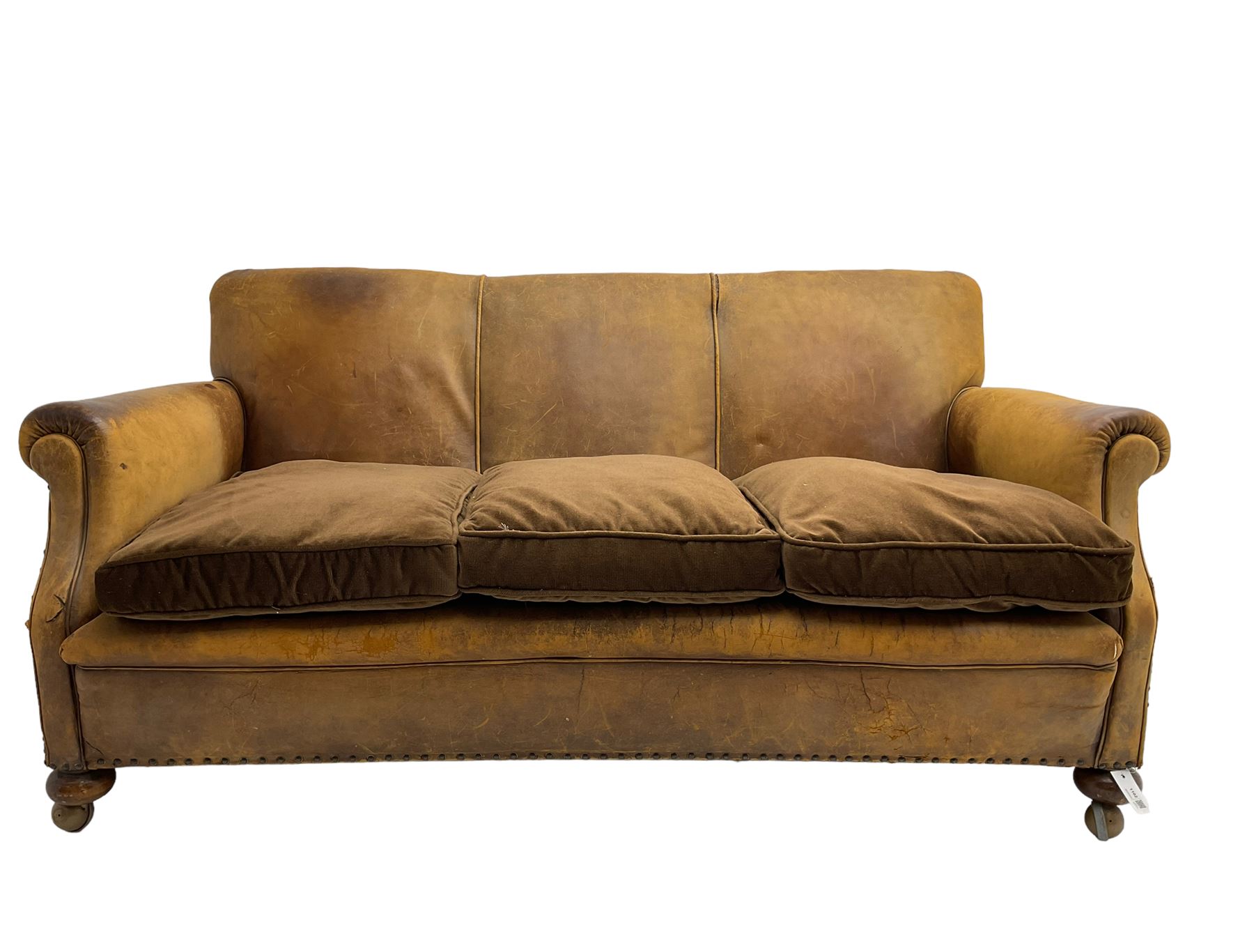 Early-to-mid-20th century three seat sofa, upholstered in worn tan leather with studwork, loose cushions upholstered in brown fabric, on compressed bun feet with castors