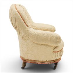 Victorian button-back nursing chair, the rounded horseshoe back enclosing a deep upholstered seat, padded arms and sprung seat in cream floral damask fabric, with crimson piping and tasselled lower fringe, on turned front legs with castors
