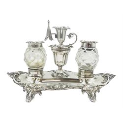 Victorian silver desk stand, of shaped form with cast acanthus leaf rim, supporting two silver mounted and faceted cut glass inkwells flanking a central pedestal cup holding a taper stick and snuffer, with engraved personal inscription to the body, the whole upon four tab feet, hallmarked Henry Wilkinson & Co, Sheffield 1856, L23.5cm, approximate weight excluding inkwells 10.41 ozt (324 grams)