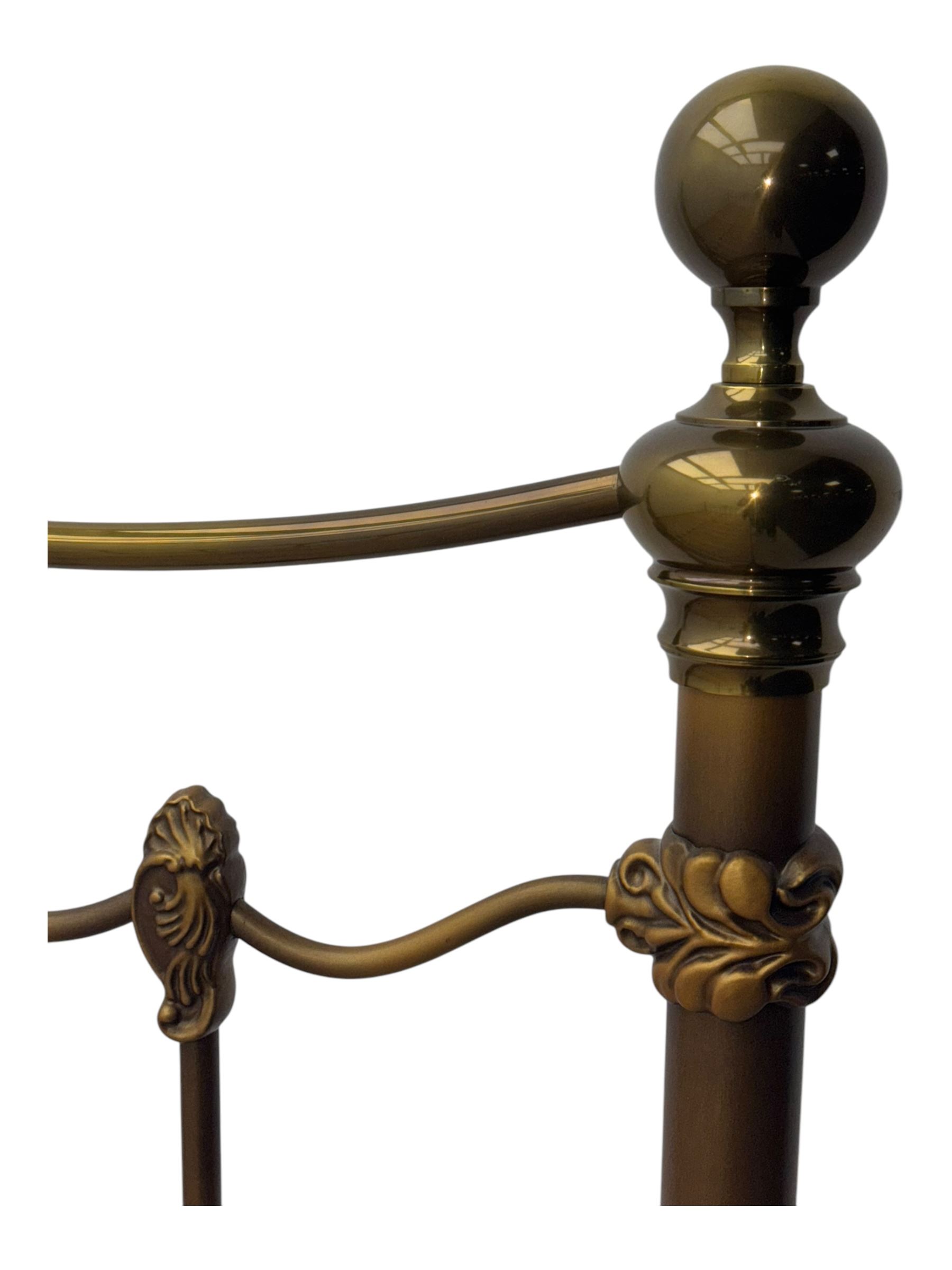 Victorian design king size brass bed frame, featuring an arched headboard and footboard with intricate cast floral and scrollwork centre motifs, flanked by cylindrical supports with ball finials and decorative collars, raised on rounded feet, with a slatted wooden base