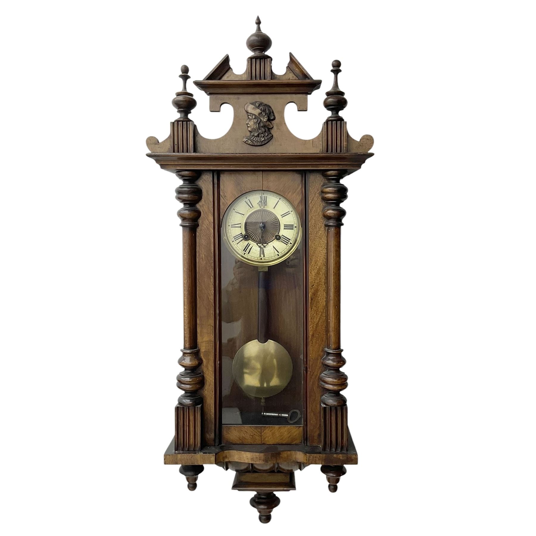 German- Late 19th century 8-day spring driven wall clock in a walnut and ebonised case, with a broken pediment and turned finials, fully glazed door with a visible pendulum and two part ivorine dial, chapter with roman numerals and minute track, rack striking movement sounding the hours and half hours on a coiled gong. With key .