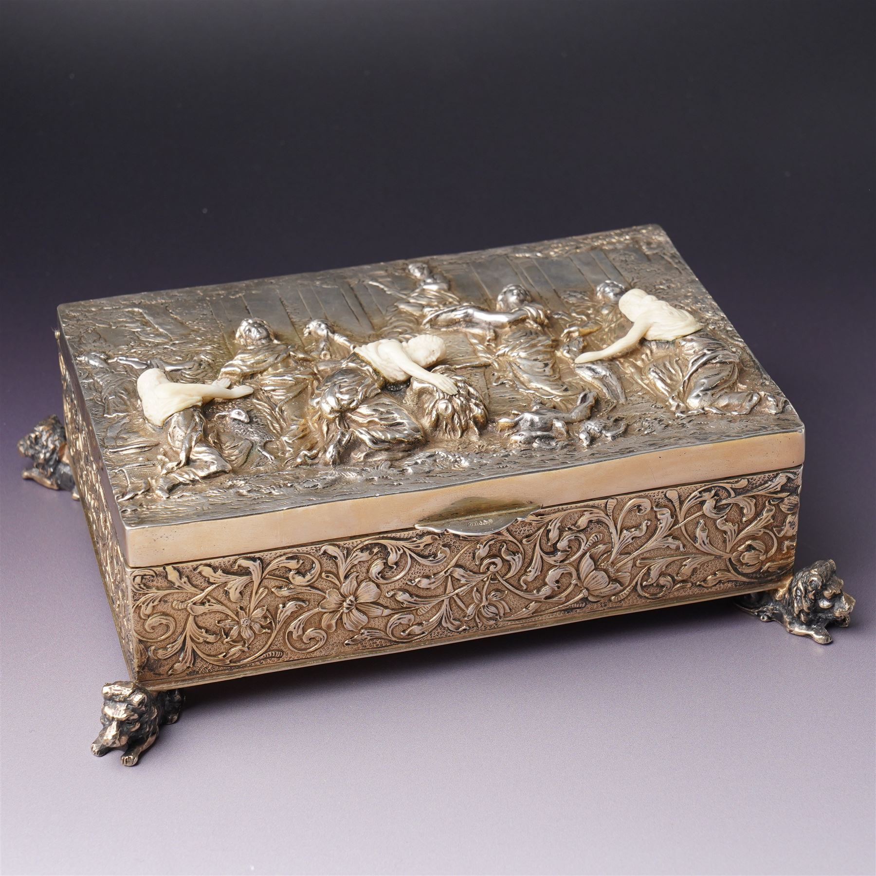 German 800 standard silver gilt and ivory mounted box, the hinged cover embossed and chased with an allegorical feasting scene, the sides with floral decoration, on four cast lion feet 21cm x 16cm x 8cm Maker Ludwig Neresheimer, Hanau circa 1900 This item has been registered for sale under Section 10 of the APHA Ivory Act