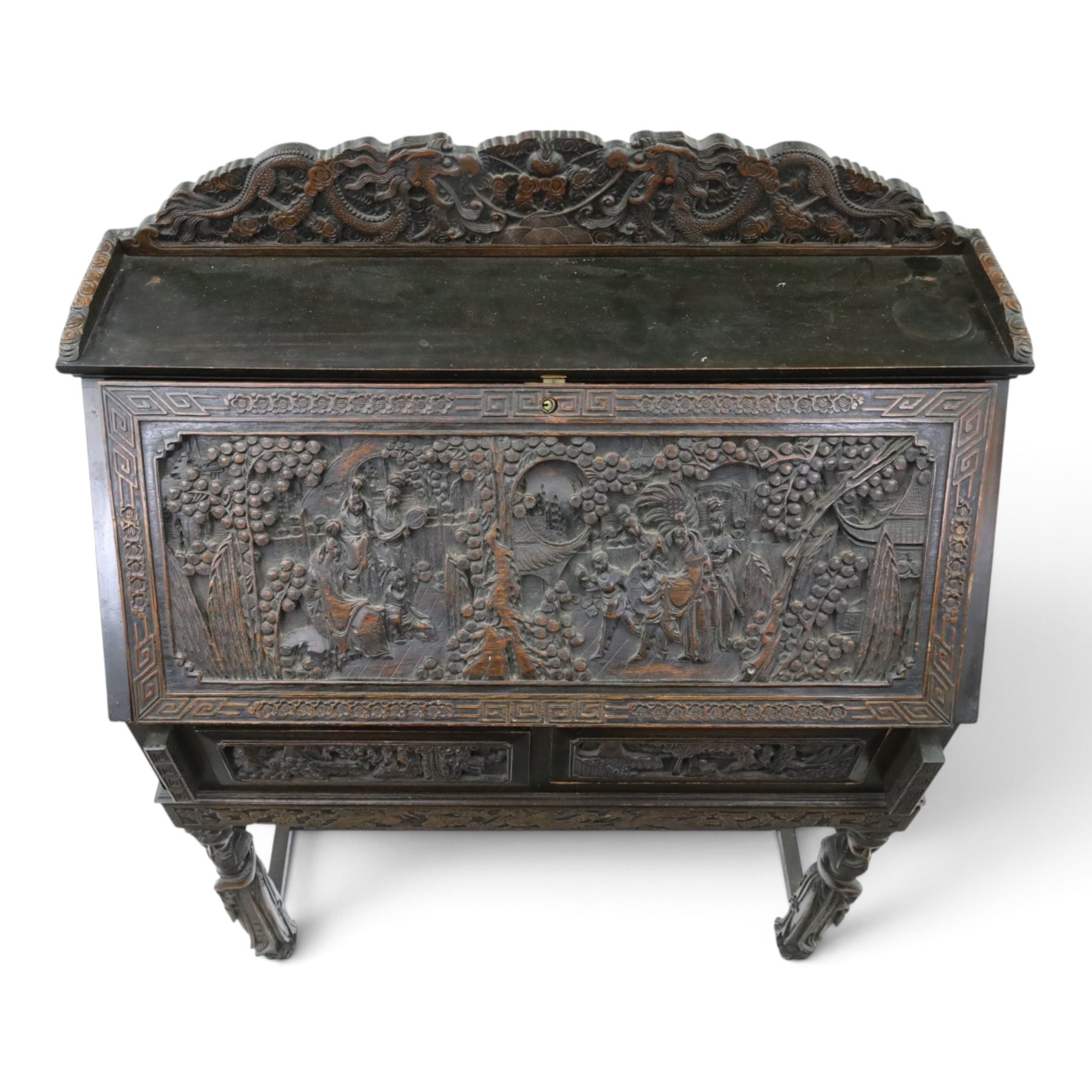 Early 20th century Chinese export carved hardwood writing bureau, the sloped fall front deeply carved with a narrative figural scene beneath pine and prunus trees, above a pair of rectangular carved panels, the shaped crest rail with entwined dragons, interior fitted with pigeonholes and a central drawer, on figural front supports with shaped stretcher