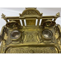 Brass desk stand, ornately embossed with floral and scrolling decoration, containing two inkwells and pen tray, flanked by a griffin to either side, together with a brass letter rack, with pierced green man mask decoration, desk stand H17cm, W30.5cm