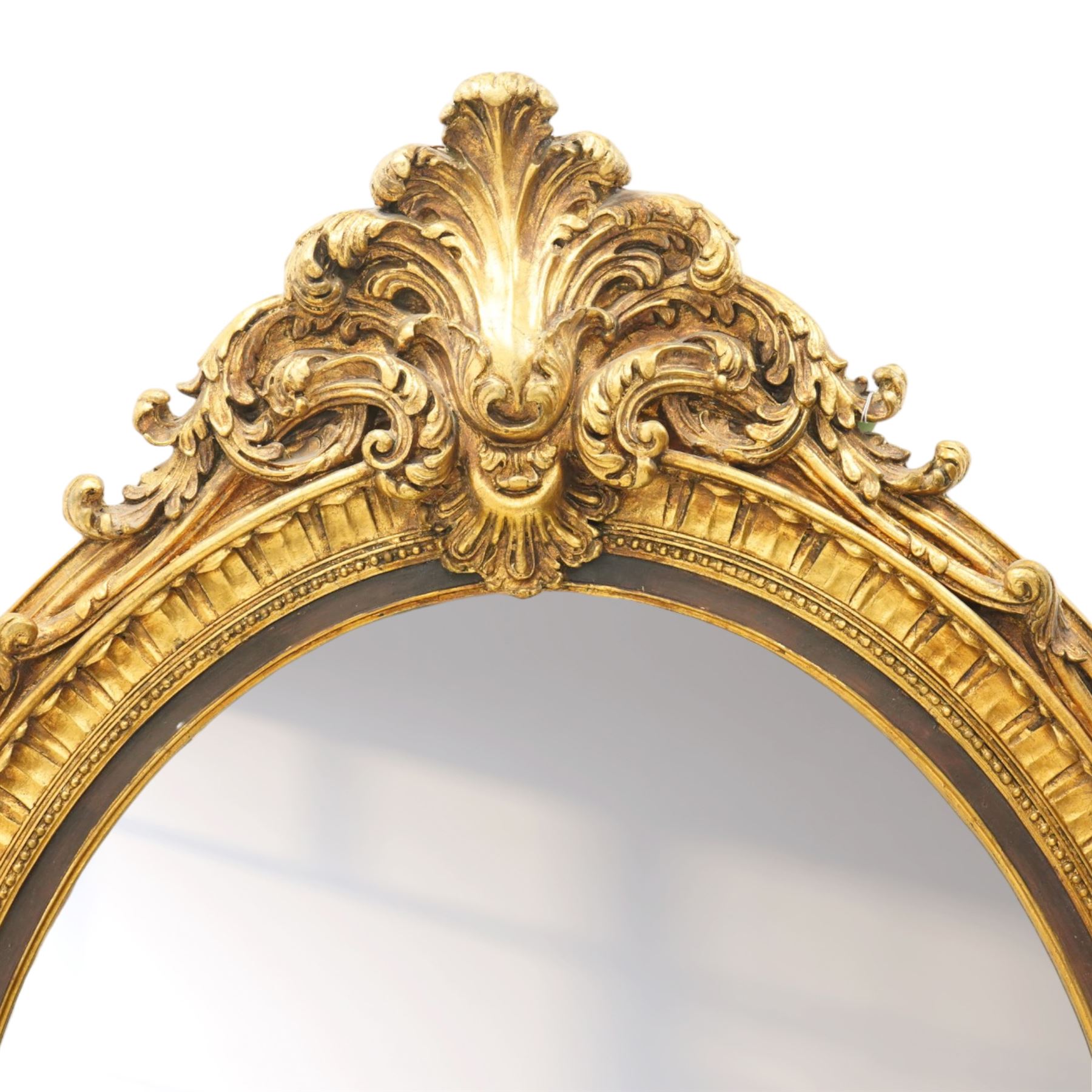 Pair of Rococo Revival gilt framed oval wall mirrors, the exuberant pediment formed of scrolling acanthus leaves and rocaille ornament, the oval bevelled plate set within with ebonised slip with outer repeating lappet decoration, over a rocaille cartouche terminal