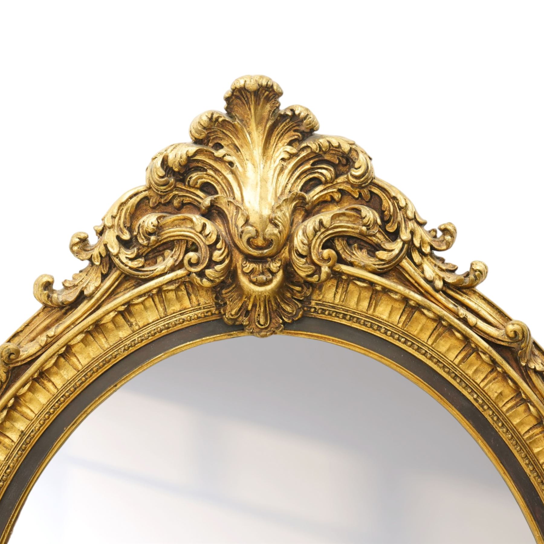 Pair of Rococo Revival gilt framed oval wall mirrors, the exuberant pediment formed of scrolling acanthus leaves and rocaille ornament, the oval bevelled plate set within with ebonised slip with outer repeating lappet decoration, over a rocaille cartouche terminal