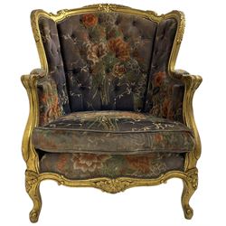 Pair of Louis XVI design gilt framed wingback armchairs, cresting rail carved and moulded with foliate decoration and C-scrolls, upholstered in buttoned floral patterned mauve velvet with loose seat cushion, scallop carved apron over cabriole supports