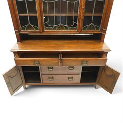 Arts and Crafts oak dresser, projecting moulded cornice above three glazed panels with coloured leaded glass floral panels, flanked by two glazed doors enclosing two adjustable shelves, open mirrored recess below with mirrored back, the base fitted with two short drawers over two central drawers flanked by carved panelled cupboard doors, ornate brass handles, raised on tapering square legs