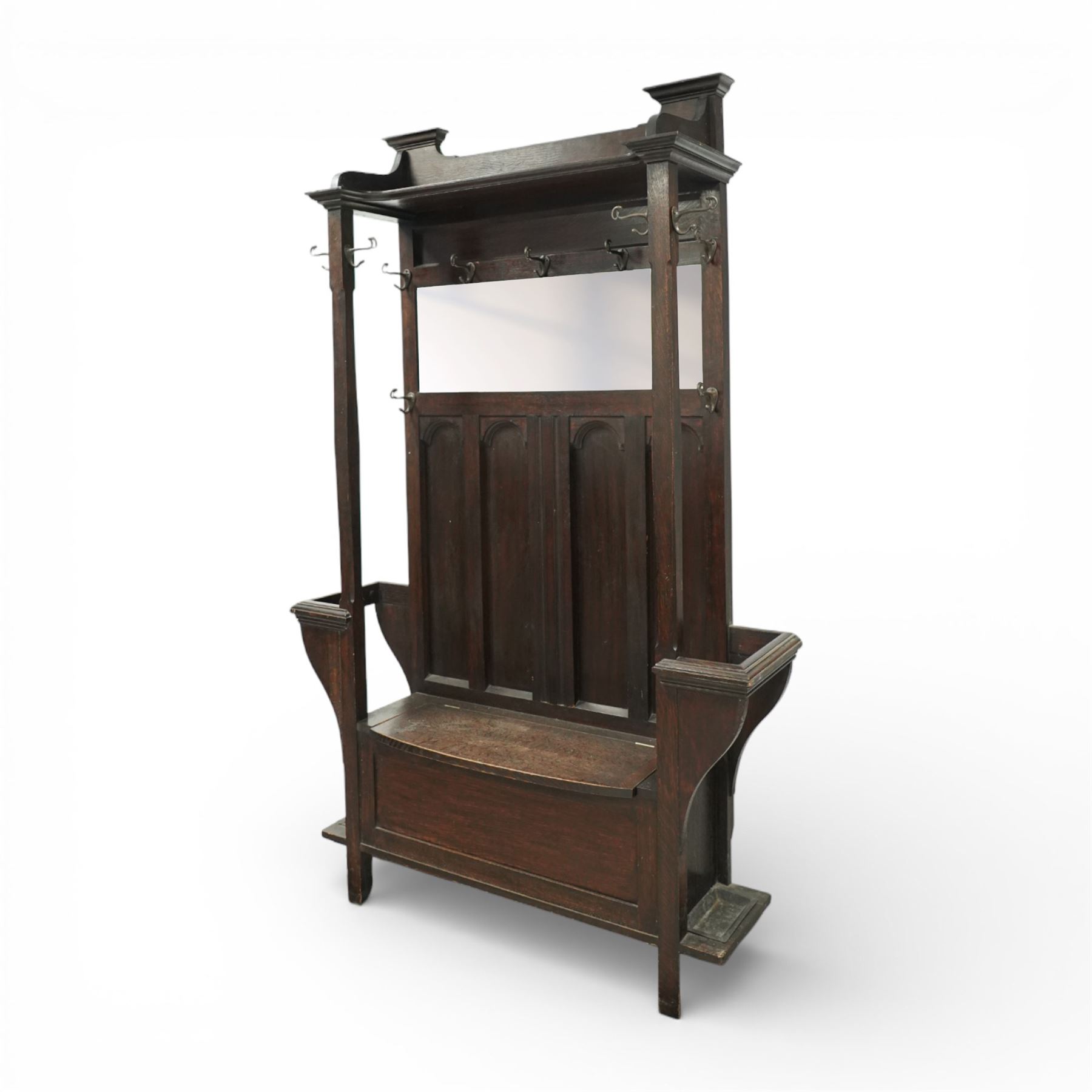 Arts and Crafts design oak settle, with projecting moulded cornice over a boarded back fitted with a rectangular bevelled mirror plate and a row of wrought iron coat hooks, the panelled back with arched fielded panels, flanked by shaped umbrella or stick stands with removable drip trays, the plank hinged seat over a panelled box base, on square section supports