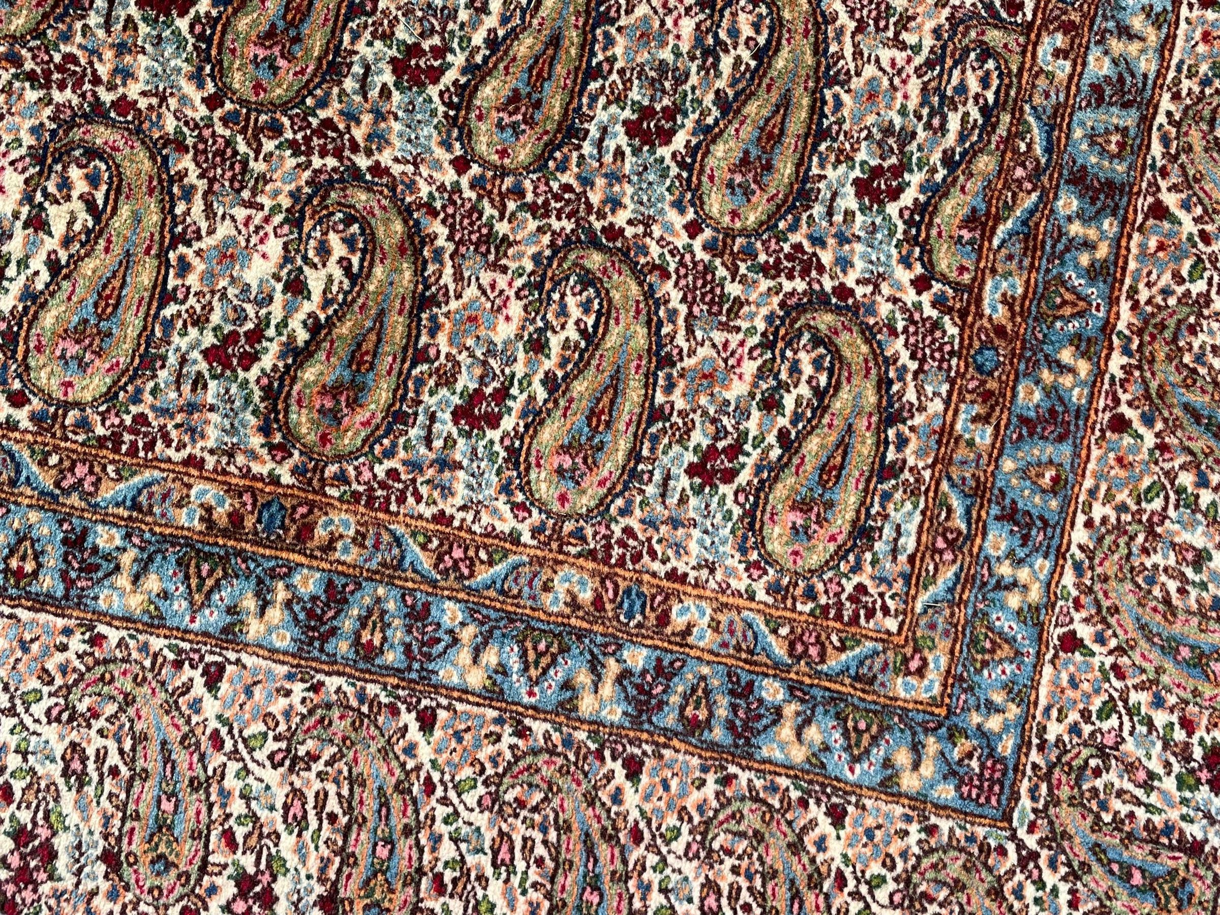 Persian Kirman carpet, the ivory ground field and border decorated with repeating Boteh motifs, interspaced with all-over floral design, blue ground border guards, the top border with signature panel