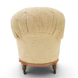 Victorian button-back nursing chair, the rounded horseshoe back enclosing a deep upholstered seat, padded arms and sprung seat in cream floral damask fabric, with crimson piping and tasselled lower fringe, on turned front legs with castors