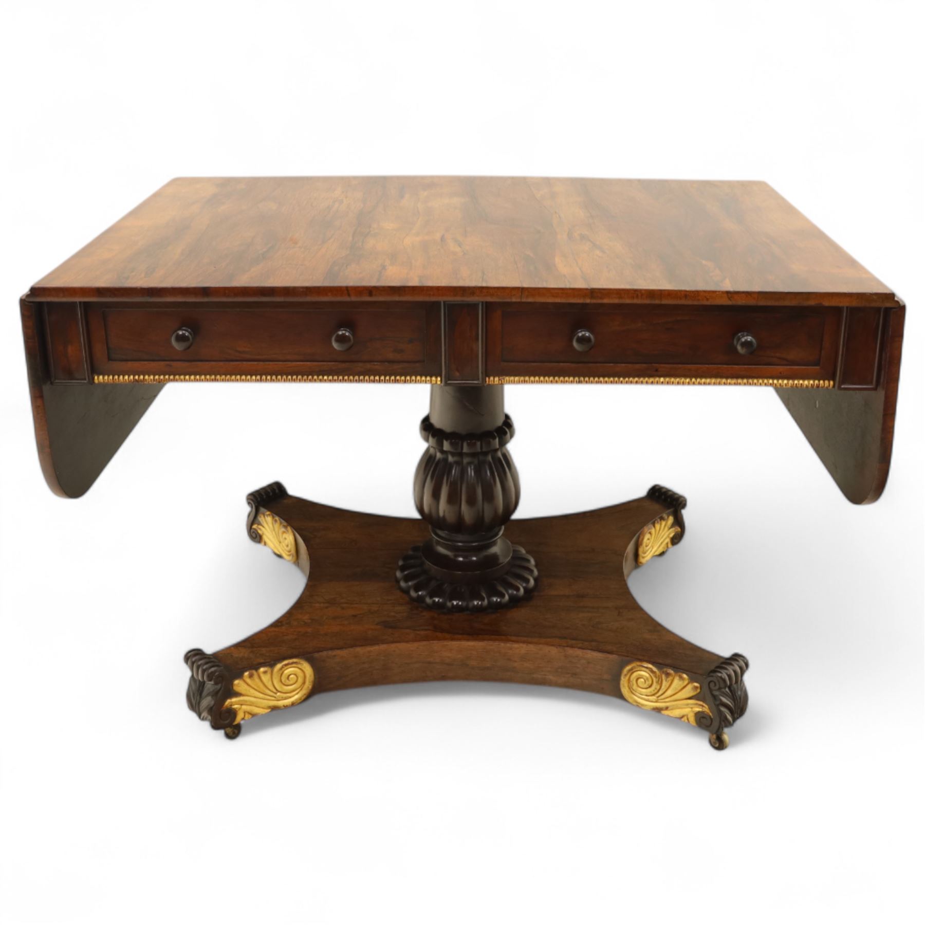Regency rosewood sofa table, rectangular top with variegated rosewood veneers, hinged D-shaped leaves, over a set of dummy drawers to the back and full-size drawers to the front, with turned knob handles and gilt beaded moulding on frieze above elegant turned, tapering, and fluted, pedestal with a well-carved collar, supported on a large, tripartite concave platform base terminating in richly carved lion paw feet with gilded acanthus leaf carving, on concealed brass castors, circa 1815, length extended 161cm 
