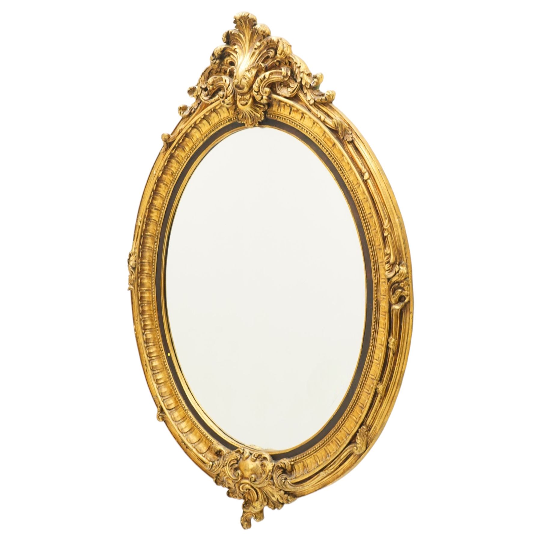 Pair of Rococo Revival gilt framed oval wall mirrors, the exuberant pediment formed of scrolling acanthus leaves and rocaille ornament, the oval bevelled plate set within with ebonised slip with outer repeating lappet decoration, over a rocaille cartouche terminal